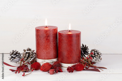 Burning red Christmas candles on a rustic white wooden background with natural potpourri elements and pine cones. 