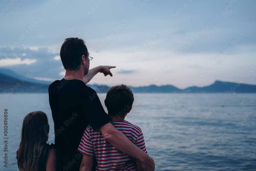 Father with son and daughter near river at sunset. Dad and kids watching sunrise together. Happy man and children relaxing on beach, family enjoying beautiful view. Bonding and togetherness