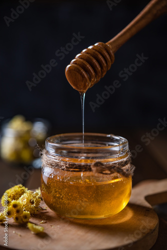 Natural floral honey drains to jar on wood background. Front view
