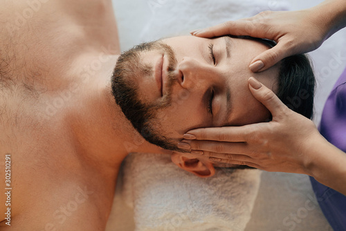 Handsome man relaxing with eyes closed during head massage at wellness resort. Massage head pain treatment
