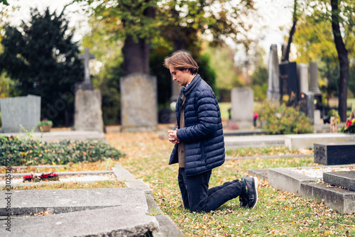 Mourning young man kneeling in front of a grave on a cemetery