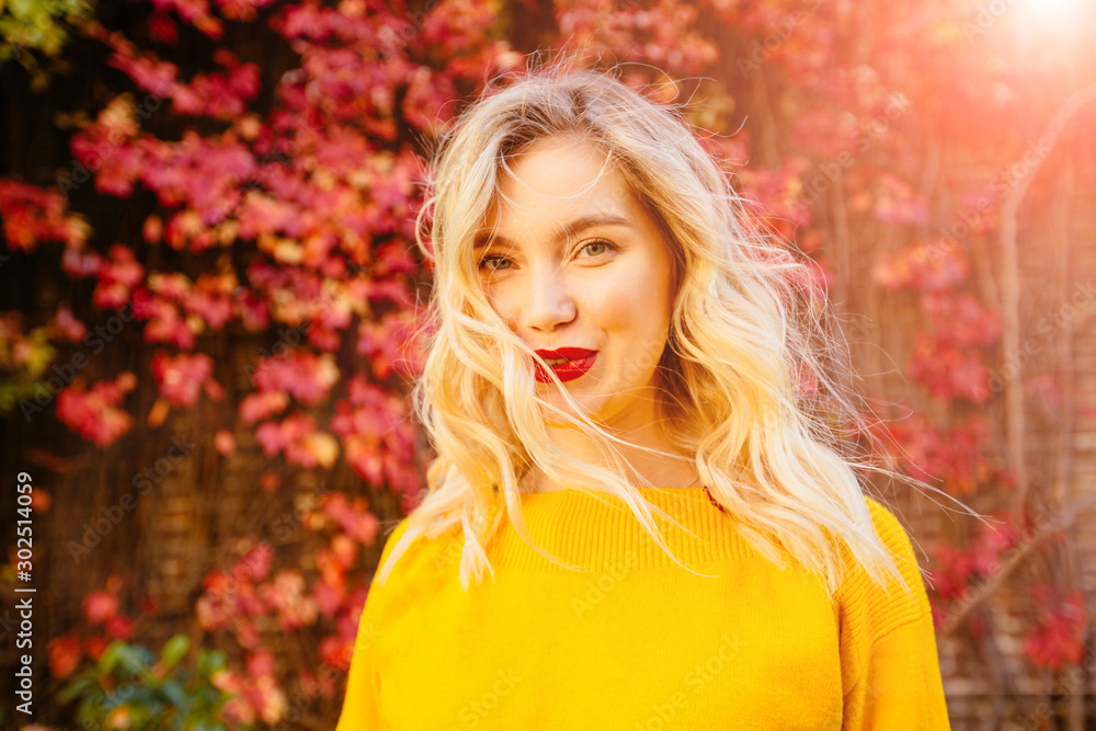 Lifestyle portrait of happy blond woman in yellow sweater with red lips flying hair, windy autumn sunny day on red brick wall with climbing grapes on background. Close up portrait. Lifestyle portrait of happy blond woman in yellow sweater with red lips flying hair, windy autumn sunny day on red brick wall with climbing grapes on background. Close up portrait.