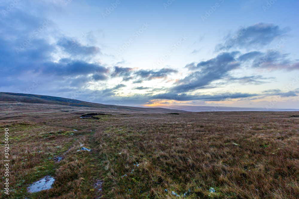 A view of grassy mountain slope at dawn with majestic orange sky in the background over mountain range summits