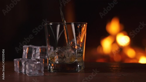 Barman pours whiskey, scotch or bourbon in a glass with ice standing on a table. Background of a fireplace with flame. Alcoholic drink concept. Fire background. Drink alone. Macro Close up Slow motion