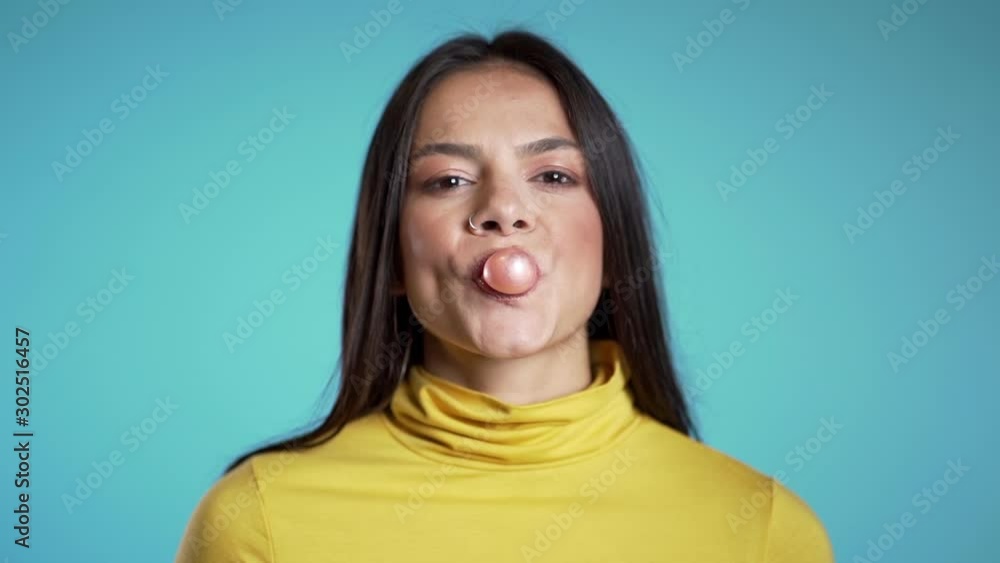 Playful girl in yellow wear blows bubblegum, chewing gum in slow motion. Pretty hispanic woman, piercing stands on blue background. Playful girl in yellow wear blows bubblegum, chewing gum in slow motion. Pretty hispanic woman, piercing stands on blue background.