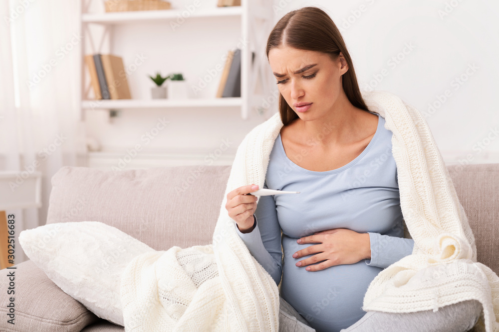 Pregnant Girl Holding Thermometer Sitting On Sofa At Home