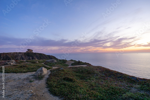 Wide-angle panorama of a beautiful lilac sunset overlooking the ocean with rocks in the foreground and a path leading up to a bunker.