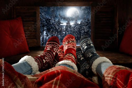 Wooden winter background of free space.Christmas december time.Woman legs with woolen socks and night landscape of forest with mountains and big moon on sky.Retro old frame and pillows decoration.