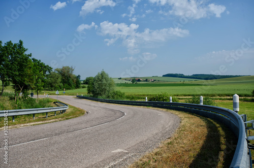 Turn view of an asphalt road. There are metal fences. In the background is a rural landscape. Far visible buildings. France. Background.