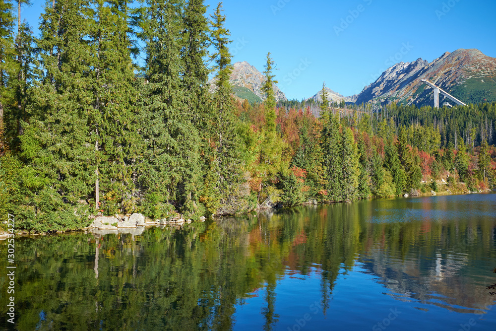 Obraz premium The beautiful Strbske pleso with reflection of the autumn park on the water. Vivid color foliage, ski jump and peaks in the background on a sunny day with clear blue sky.