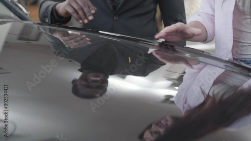 View of new black car hood with reflection of two people. Young Caucasian woman caressing automobile, smiling African American man taking her hand. Multiracial couple admiring new car in showroom.