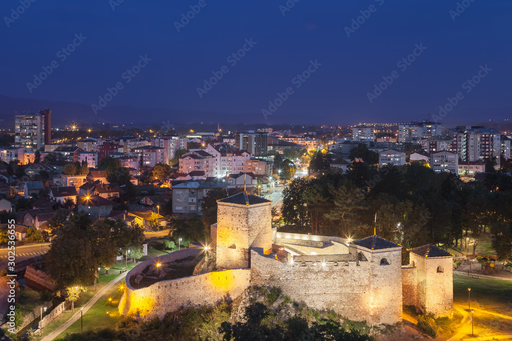 Beautiful, illuminated ancient fortress in front of the cityscape during late blue hour with city lights and lighten windows in the houses and buildings