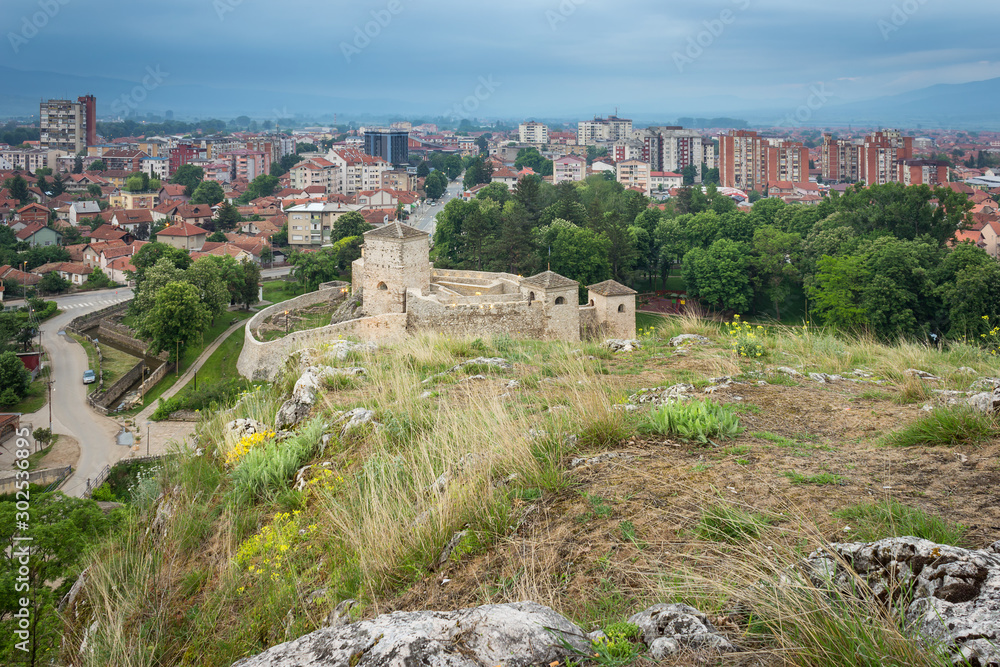 Low angle shot with foreground rocks and vivid yellow flowers in a foreground and ancient fortress and Pirot cityscape at the distance during blue hour