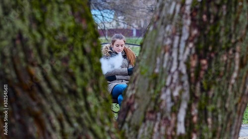 Vaping teenager. Young pretty white caucasian teenage girl in green jacket with hood smoking an electronic cigarette on the bench on the street in the autumn day. Bad habit. Vape activity.