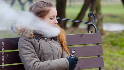 Vaping teenager. Young pretty white caucasian teenage girl in green jacket with hood smoking an electronic cigarette on the bench on the street in the autumn day. Bad habit. Vape activity.