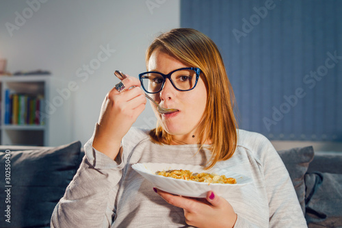 Portrait of young caucasian woman eating pasta lunch or dinner sitting on her bed sofa at home holding white plate and spoon wearing sweater and glasses photo