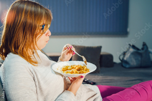 Portrait of young caucasian woman eating pasta lunch or dinner sitting on her bed sofa at home holding white plate and spoon wearing sweater and glasses