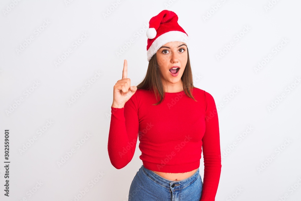 Young beautiful girl wearing Christmas Santa hat standing over isolated white background pointing finger up with successful idea. Exited and happy. Number one. Young beautiful girl wearing Christmas Santa hat standing over isolated white background pointing finger up with successful idea. Exited and happy. Number one.