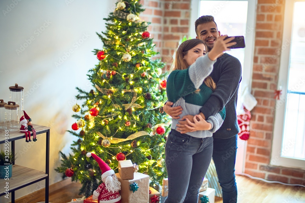 Young beautiful couple smiling happy and confident. Standing and hugging make selfie by camera around christmas tree at home Young beautiful couple smiling happy and confident. Standing and hugging make selfie by camera around christmas tree at home