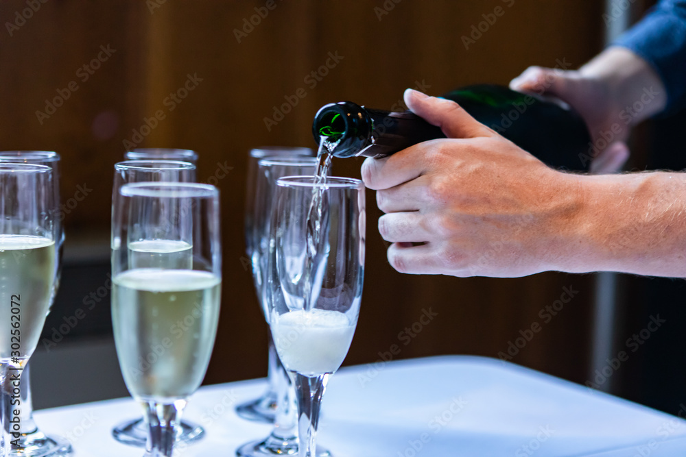 Close up photo of a person pouring in wine and champagne in empty crystal glasses that are standing on a white clothed buffet table 