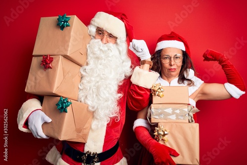 Middle age couple wearing Santa costume holding tower of gifts over isolated red background Strong person showing arm muscle, confident and proud of power
