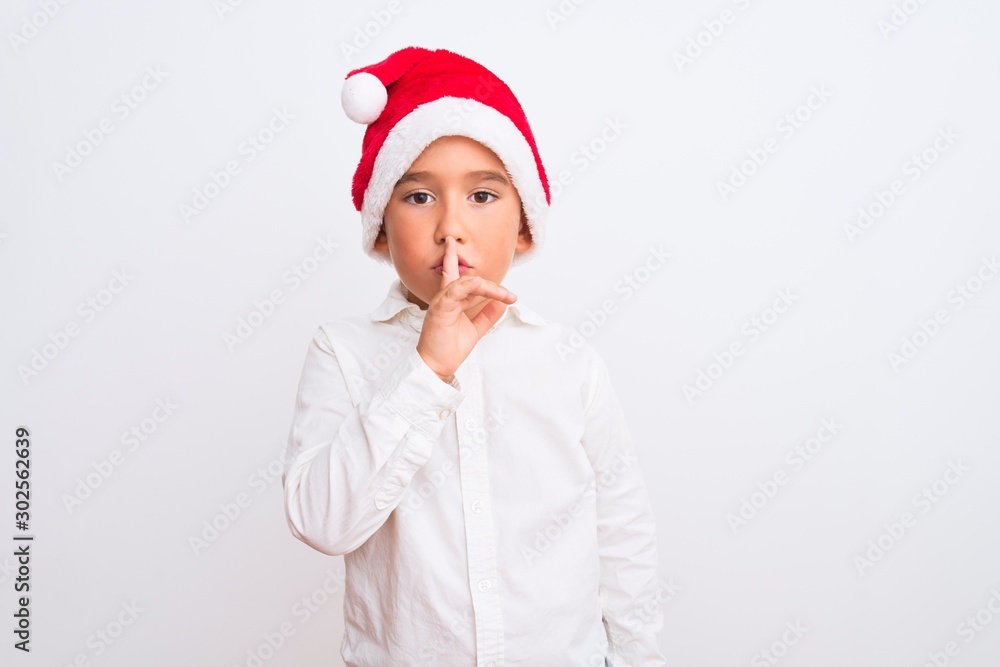 Beautiful kid boy wearing Christmas Santa hat standing over isolated white background asking to be quiet with finger on lips. Silence and secret concept. Beautiful kid boy wearing Christmas Santa hat standing over isolated white background asking to be quiet with finger on lips. Silence and secret concept.