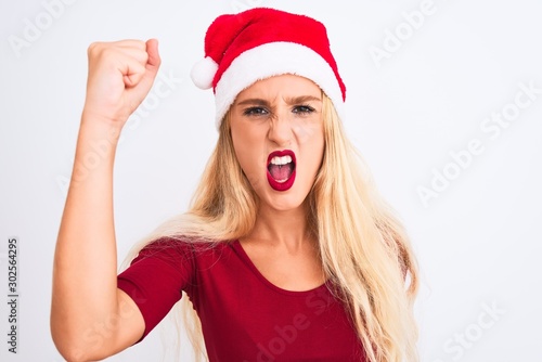 Young beautiful woman wearing Christmas Santa hat over isolated white background angry and mad raising fist frustrated and furious while shouting with anger. Rage and aggressive concept.