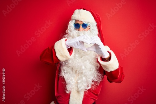 Middle age man wearing Santa Claus costume and sunglasses over isolated red background smiling in love showing heart symbol and shape with hands. Romantic concept.