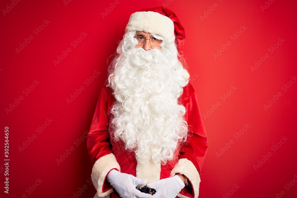 Middle age handsome man wearing Santa costume standing over isolated red background Relaxed with serious expression on face. Simple and natural looking at the camera.