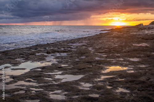 A Rocky Shore In Hawaii Looking out to the Ocean in the Evening - with a Vibrant Sunset in the Background and Reflected in Pools of Water on the Rocks