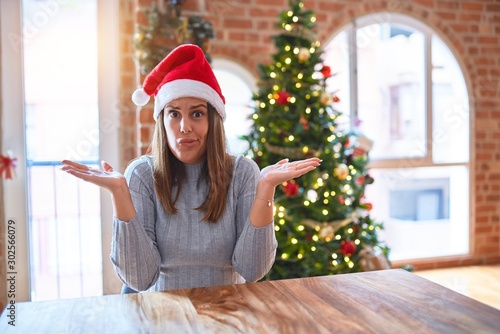 Young beautiful woman wearing santa claus hat at the table at home around christmas decoration clueless and confused expression with arms and hands raised. Doubt concept.