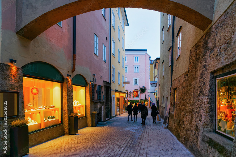 Fototapeta premium People at Narrow Street with shops and stores at Old city of Salzburg, Austria. Building architecture of Mozart town, Europe, winter. Panorama and landmark. Cityscape in Evening. Travel and tourism.