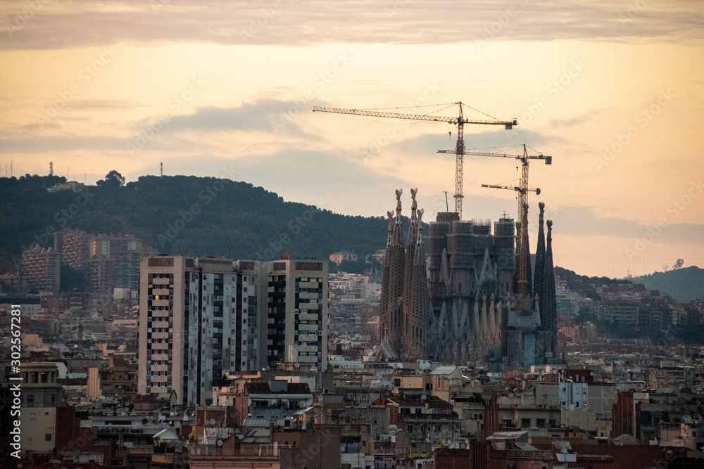 Obraz premium A view of the still under construction Sagrada Familia with its construction cranes during sunset with mountains in the background