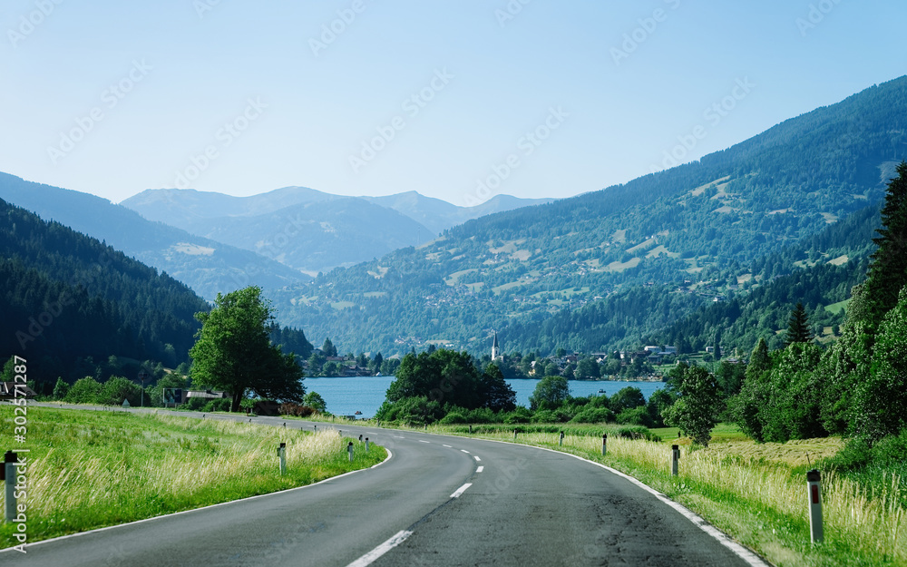 Car Road at lake Field am See of Carinthia of Austria. Landscape with motorway for rides and driving at pond and blue sky in spring or summer. Scenery in green Alps of Europe.