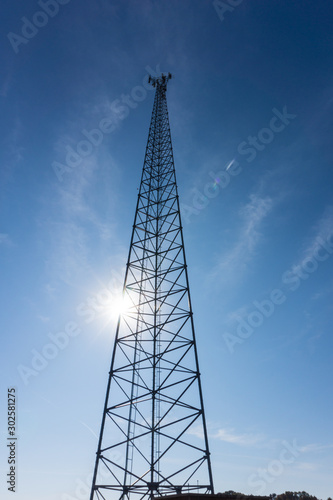 Bright sun shines behind radio tower antenna in a blue sky.