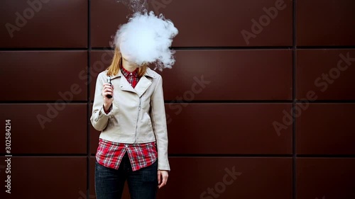 Vaping teenager. Young pretty white girl in casual clothes smoking an electronic cigarette opposite dark modern backgroun on the street in the autumn. Bad habit. Vape activity.