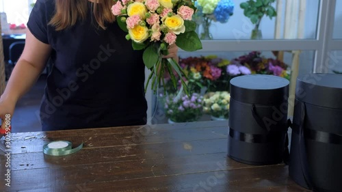 Floral business concept. Professional woman florist works makes bouquet ties roses stems with cutted green ribbon at table in flower shop, hands closeup. Creates bunch composition in floristic store.