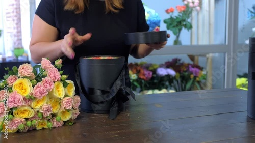 Creates bunch composition in floristic studio store. Professional woman florist makes creates bouquet with roses preparing black packaging box in shop, hands closeup. Floral business concept.