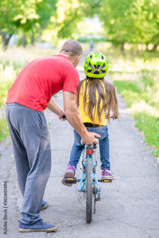 Fototapeta premium Active family leisure. Father teaches his daughter to ride a bicycle in the summer park. Back view