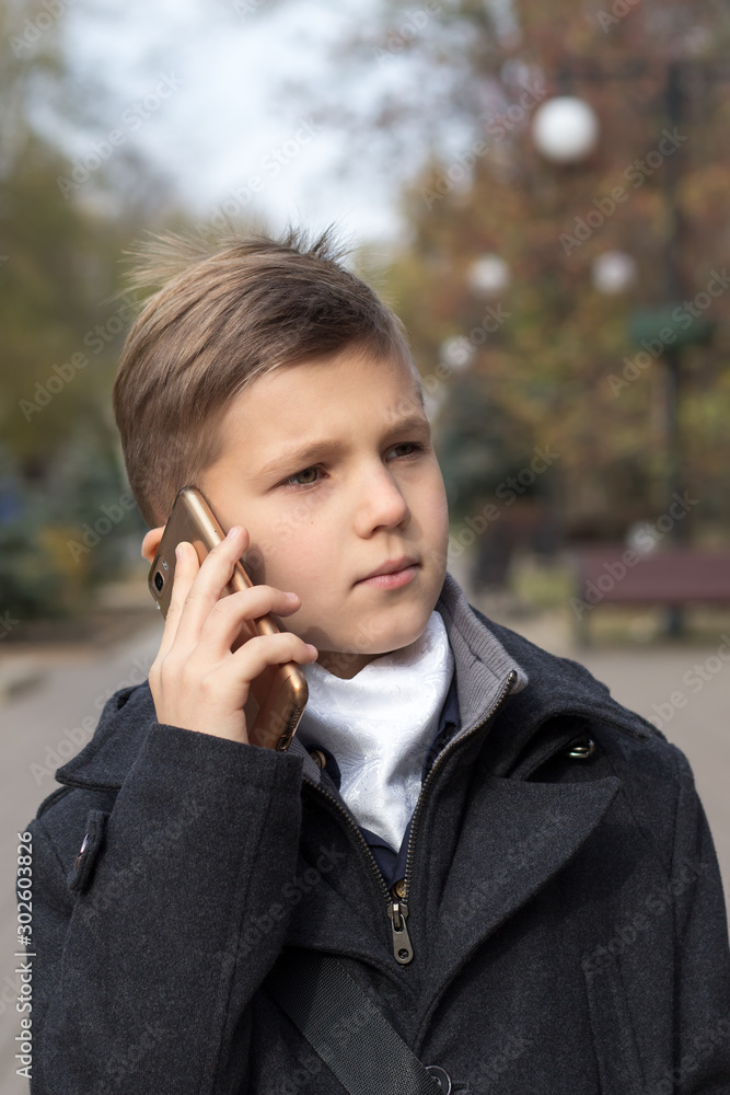 Portrait of a schoolboy dressed in a business suit talking on a cell phone, holding it in his hand near his ear. Front view with a serious expression and a slight smile. Autumn day in the boulevard Portrait of a schoolboy dressed in a business suit talking on a cell phone, holding it in his hand near his ear. Front view with a serious expression and a slight smile. Autumn day in the boulevard
