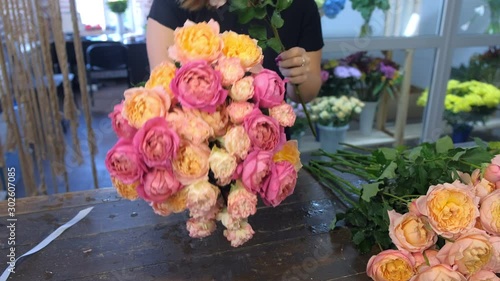 Florist woman making bouquet of pink and orange peonies in flower shop, closeup hands. Floral business concept. She creates professional bunch composition in floristic studio store at table.