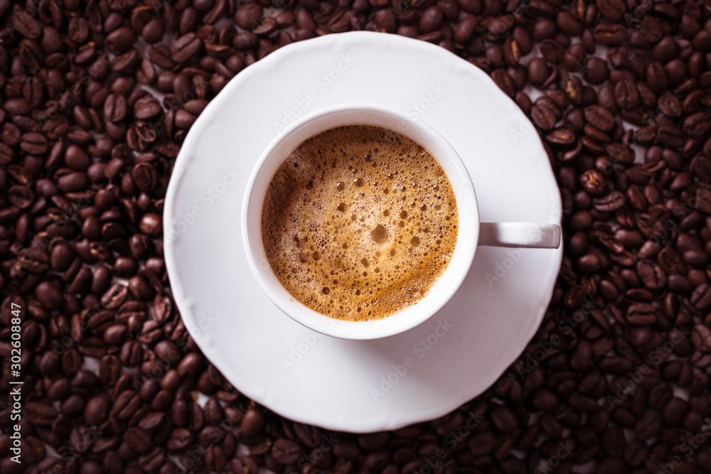 Fototapeta premium Cup of fresh americano or espresso coffee with golden foam froth on pile of brown raw coffee beans on white marble table background. Morning hot drink, coffee break, cope space