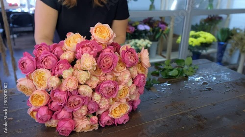 Florist woman making bouquet of pink and orange peonies in flower shop, closeup hands. Floral business concept. She creates professional bunch composition in floristic studio store at table.