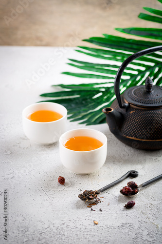 Asian style tea ceremony, black iron kettle teapot with white bowl cups with palm leaf on light background. Healthy hot detox drink, copy space
