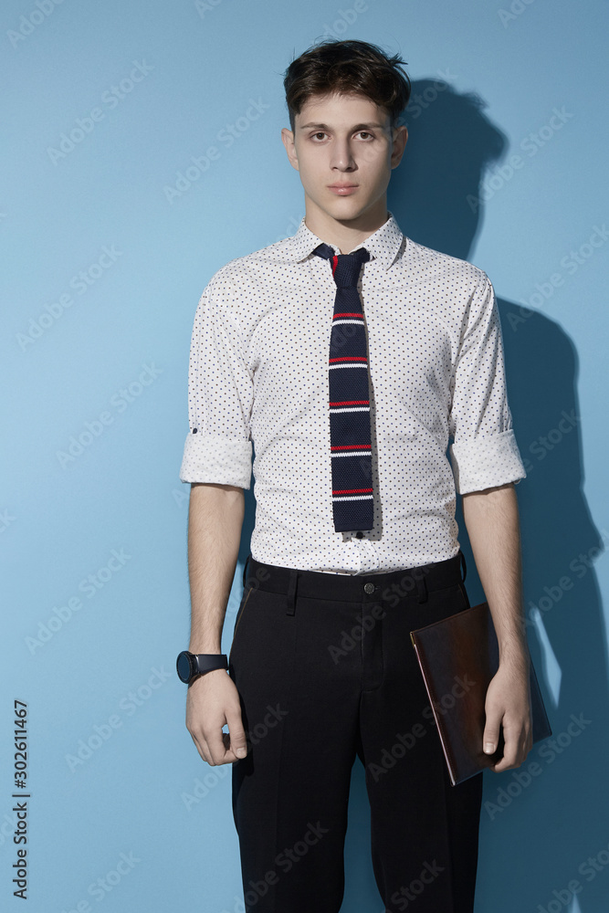 Fototapeta premium Medium full shot of a young dark haired man in a black trousers and white printed shirt with a deep blue knit tie with red and white stripes. The man with a black watch is holding a leather folder. 