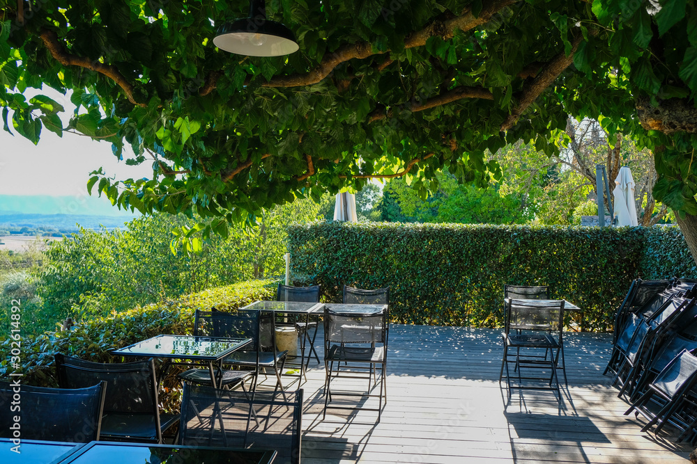 Empty glass tables on wooden terrace under magnificent tree. Deep sunshine. Provence tourism.  Joucas.