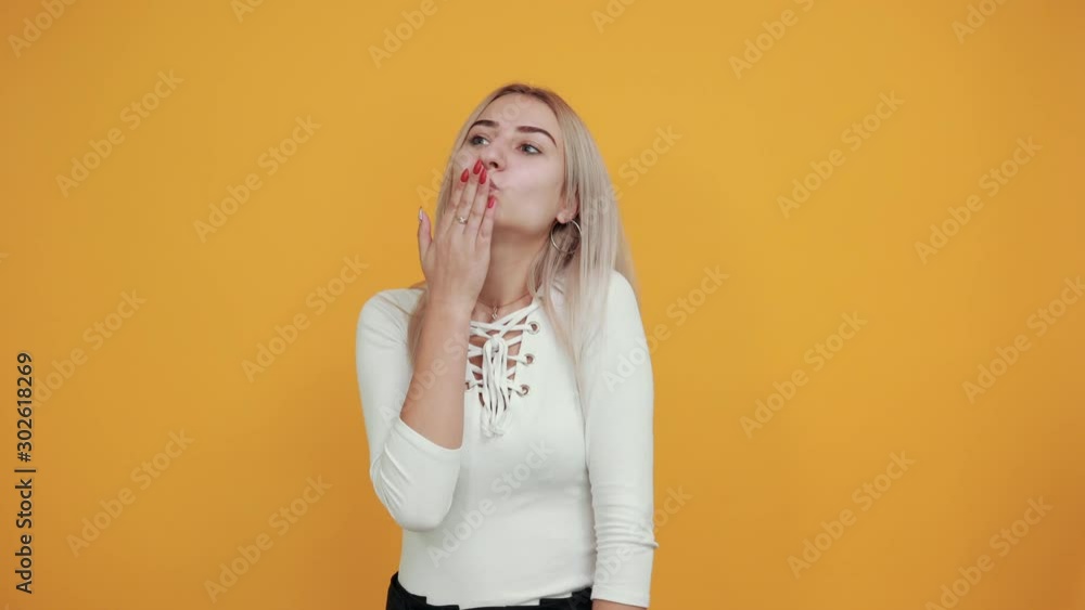 Pretty stylish young woman in white shirt send air kiss aside isolated on orange wall background studio portrait. People sincere emotions lifestyle concept. Pretty stylish young woman in white shirt send air kiss aside isolated on orange wall background studio portrait. People sincere emotions lifestyle concept.