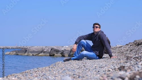 Guy on spring vacation. Happy man tourist traveller sits at stone beach and looks at sea on vacation. He is wearing sunglasses, jeans and sweatshirt. Sunny warm day at seashore. Travel journey trip. photo