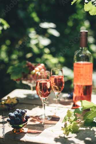 Rose wine tasting, glass of rose wine poured from bottle outdoors in garden party in vineyard, ripe grapes on wooden table, sunlight, harvest time, copy space