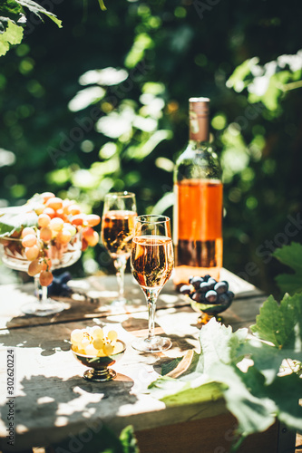 Rose wine tasting, glass of rose wine poured from bottle outdoors in garden party in vineyard, ripe grapes on wooden table, sunlight, harvest time, copy space